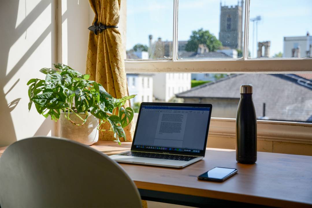 Laptop, mobile phone and water bottle on a desk with a view of a city outside a window