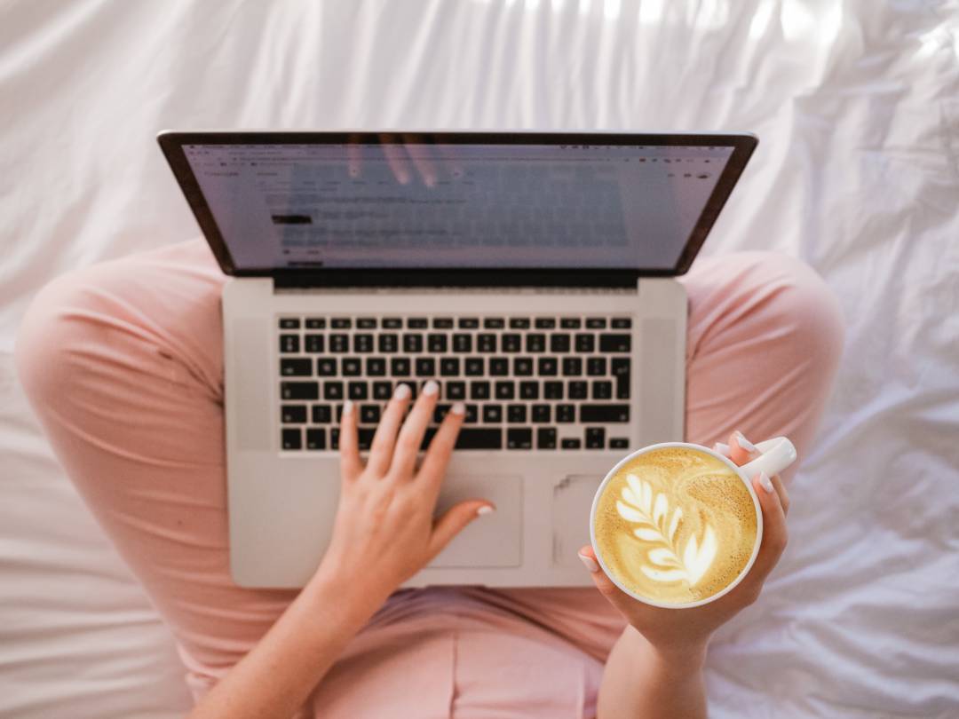 Person sitting in bed with a laptop and a cappuccino in hand