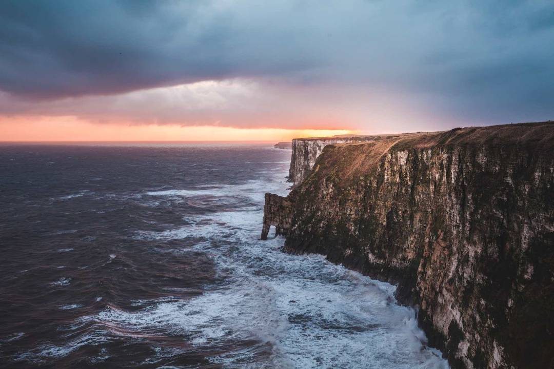 View Of Bempton Cliffs On The Right Hand Side With Cloudy Skies And Roaring Ocean On The Left