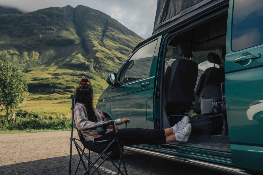 Woman On Chair Next To Green Campervan With Hill Backdrop
