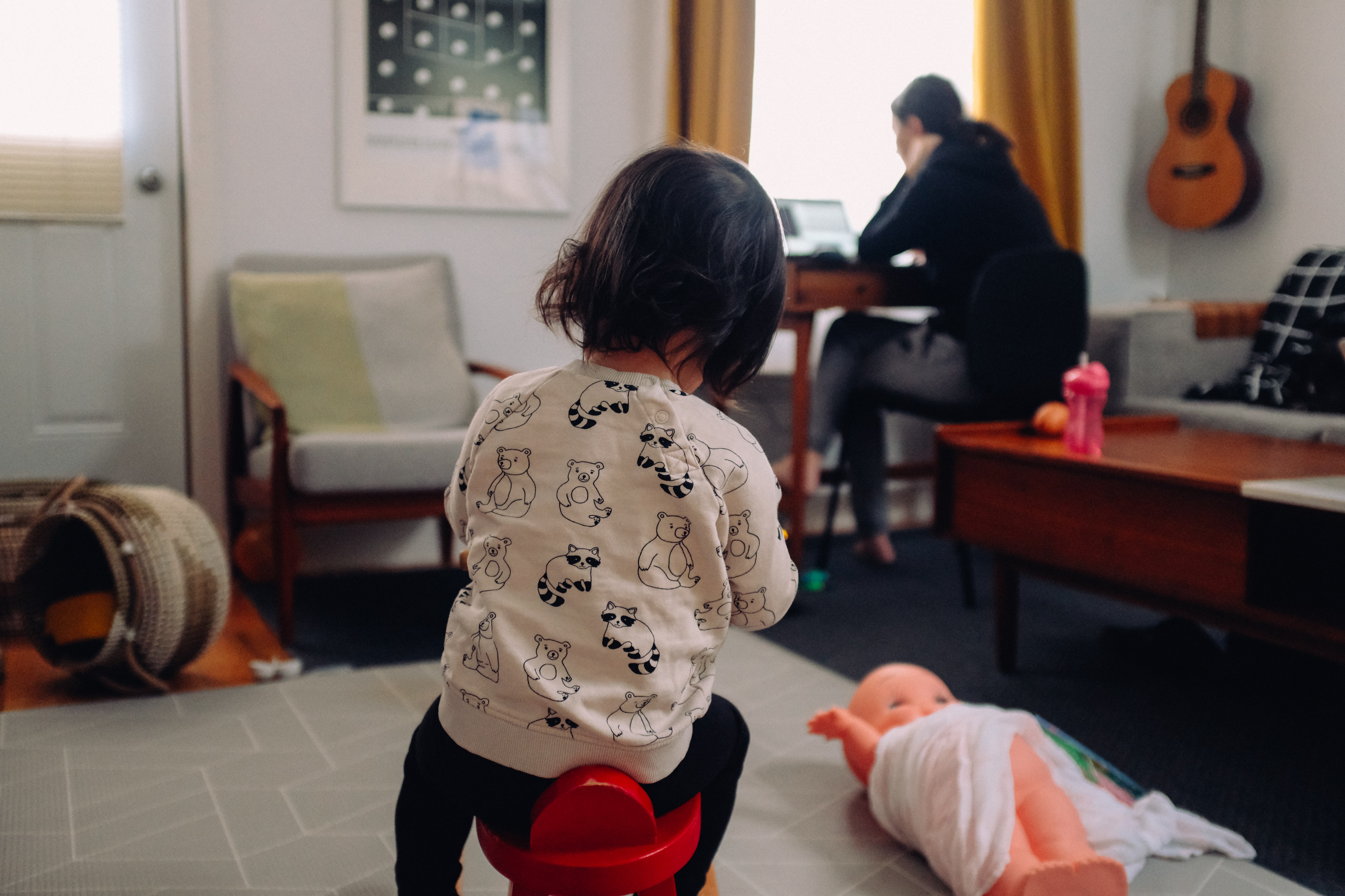Child playing in a living room with a woman working on a laptop in the background